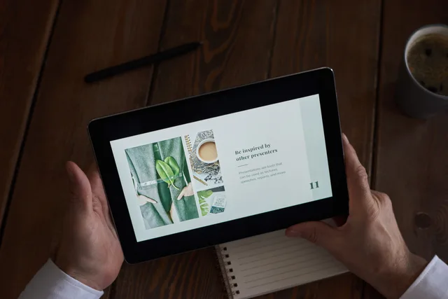 Close-up of hands holding a tablet showing a presentation on a wooden desk. Reviewing layout quality with the same source material